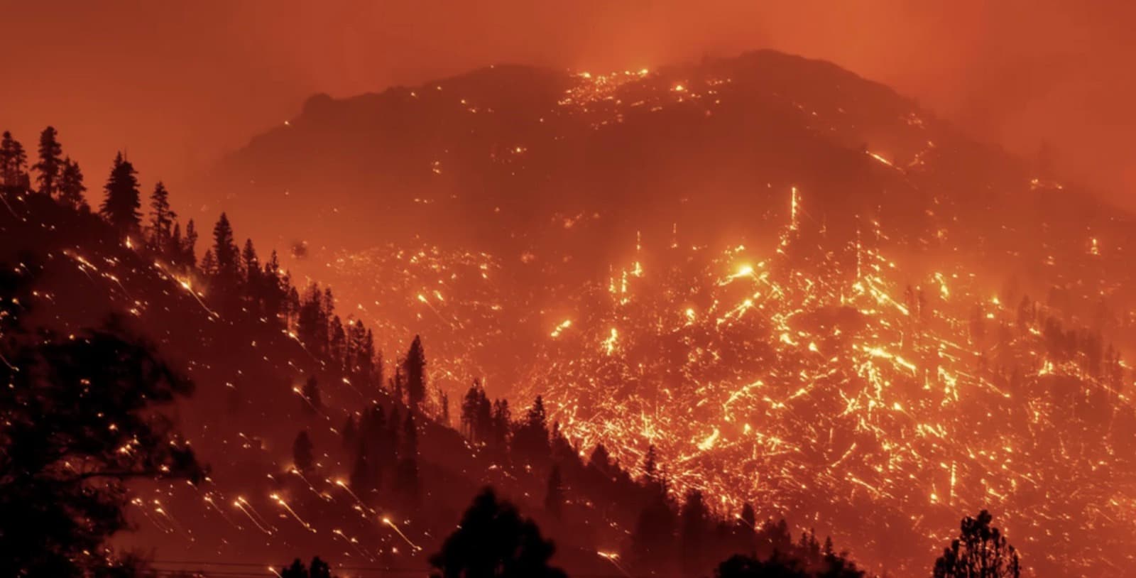An aerial night view of a mountainside wildfire, with thousands of embers and spot fires scattered across the slopes under a smoke-orange sky.